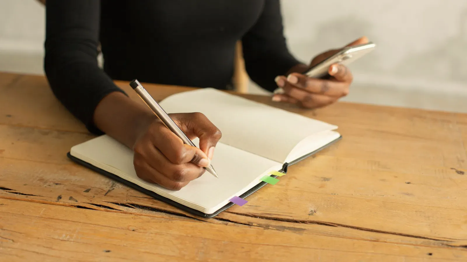 A person at a wooden table, writing in a notebook while holding a phone.
